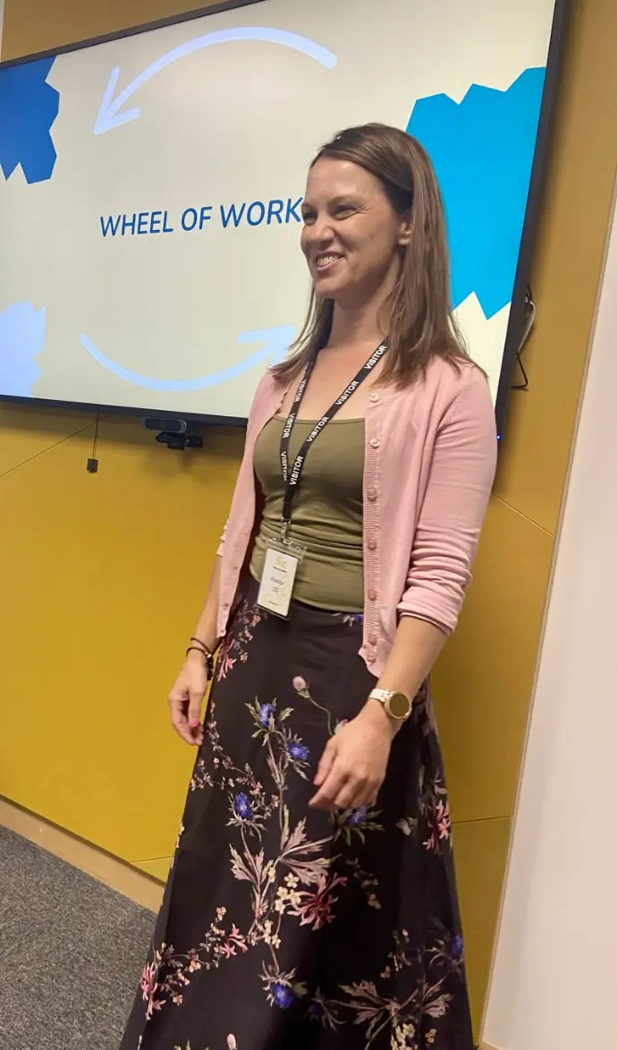 Caroline Barnes stood in fron tof a yellow wall with a screen to her right that says 'wheel of work'  She has been presenting a workshop and is smiling