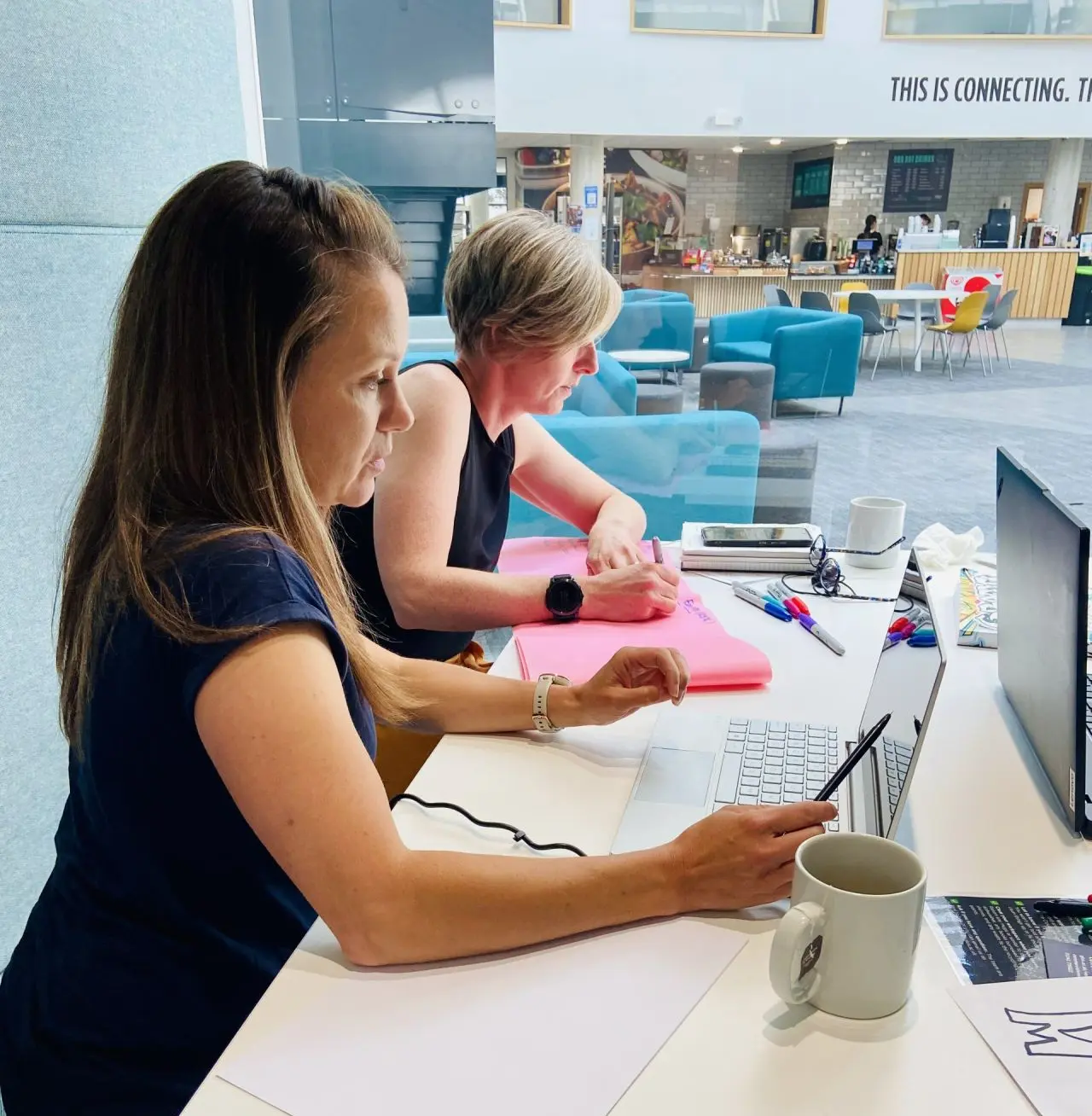 A side picture of Caroline and Faye working at a desk with sofas in the background.  Caroline is wearing a blue t shirt and is pointing to a laptop with a pen in her hand.  Faye is writing with a marker pen on a large piece of pink paper.  There are pens and a coffee cup on the desk also