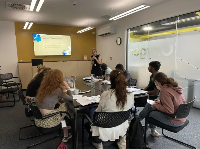 A training room with 8 people sat around a rectangular table facing the front of the room.  Faye Ingram is standing at the front of the room talking to the group with a screen to her right.  She is wearing a black dress