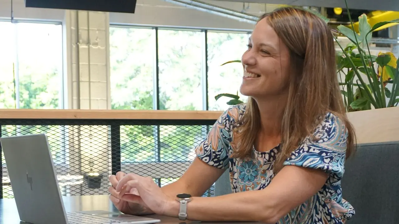 Caroline barnes is sat on the right of the image looking towards the left and smiling.  She is at a table and has a laptop open in front of her.  She is wearing a floral shirt an smiling