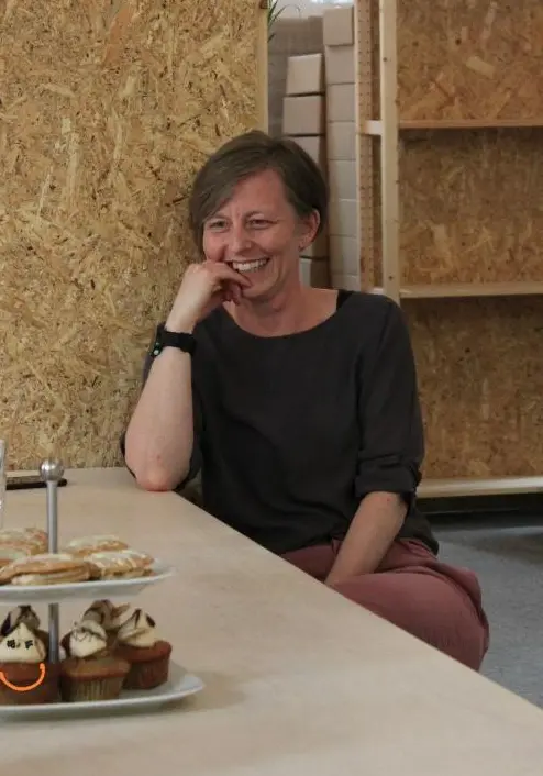 Faye Ingram is sat at a table wearing a black top.  She has her chin resting on her hand and is  looking at a cake stand on the table and smiling.  She is surrounded by wooden shelving