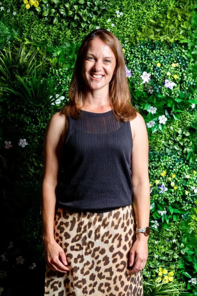 Caroline Barnes is dressed in a black vest and leopard print skirt .  She is stood in front of a bright green leaf display and is facig the camera smiling