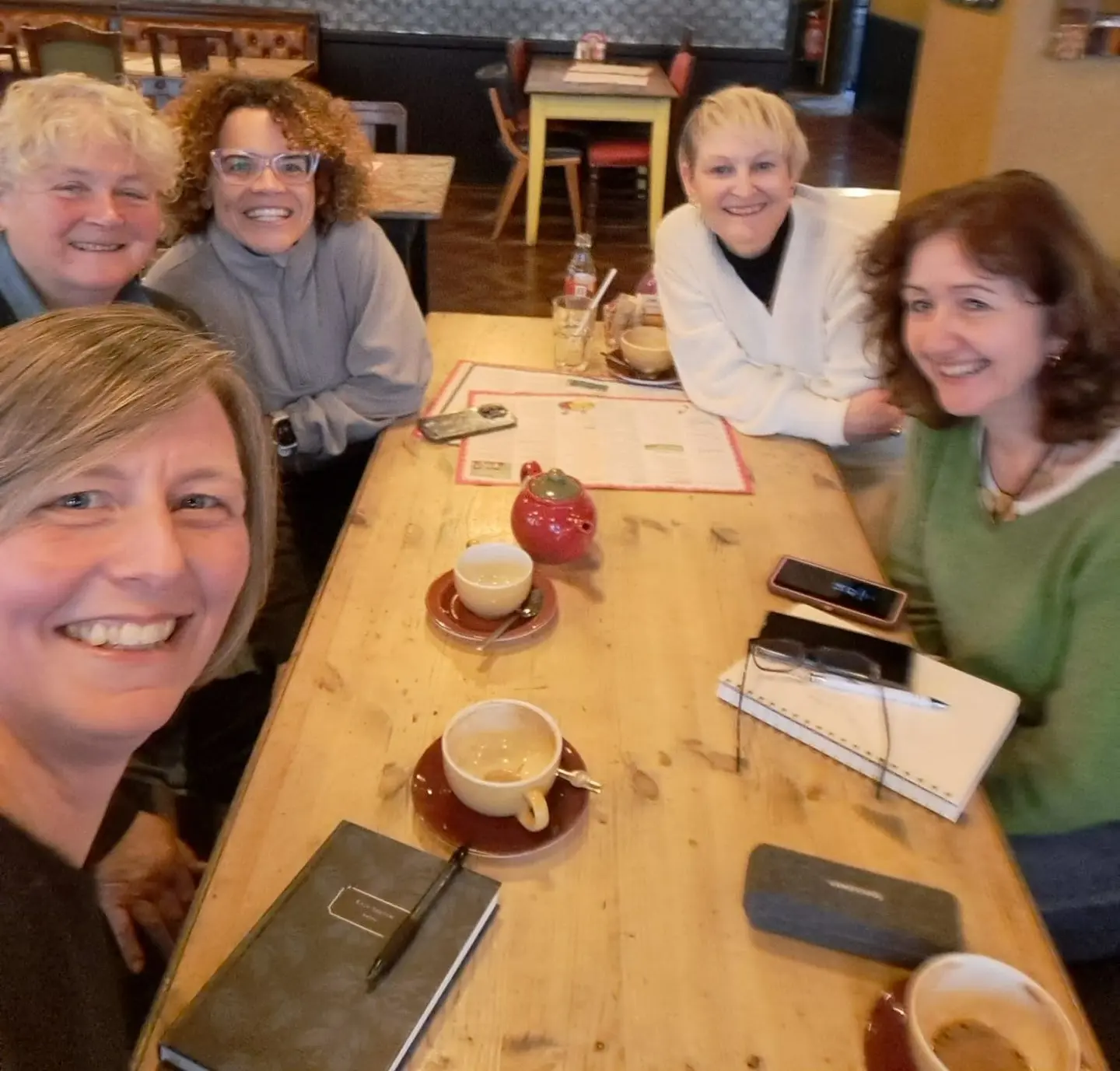 Faye Ingram is sat with a group of 4 other ladies around a table.  They are all looking towards the camers and smiling.  There are coffee cups and notebooks on the table in front of them
