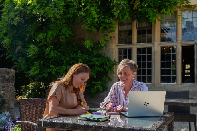Caroline Barnes and Faye Ingram are sat outside of a window at a table working at a laptop and writing some ideas down