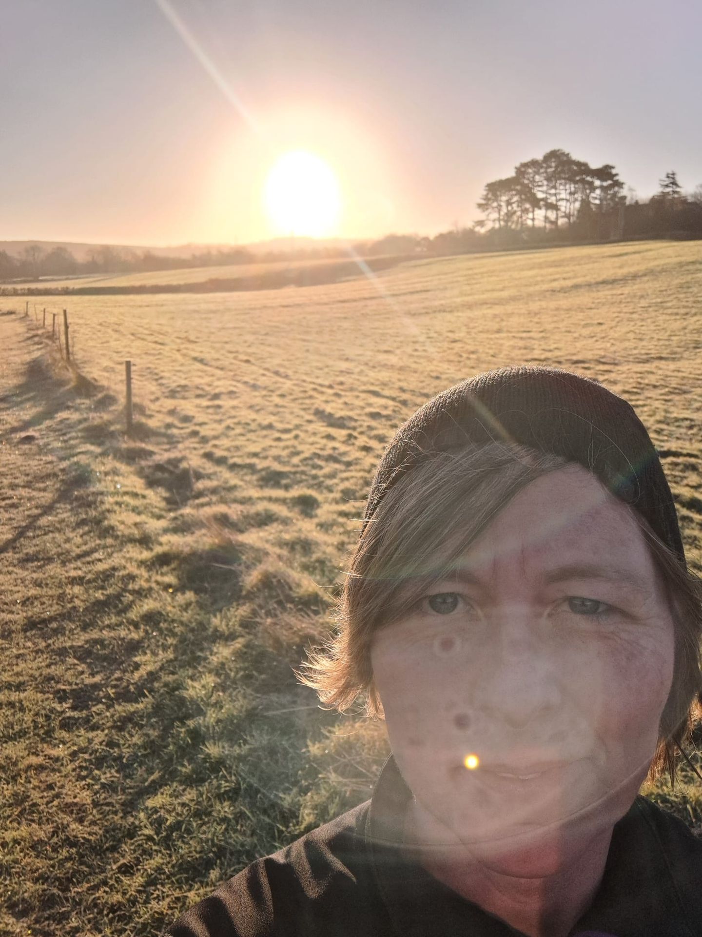 Selfie picture of Faye in her running kit in the forefront of the picture.  In the background is a field with morning dew and the sun rising above the tree line in the background